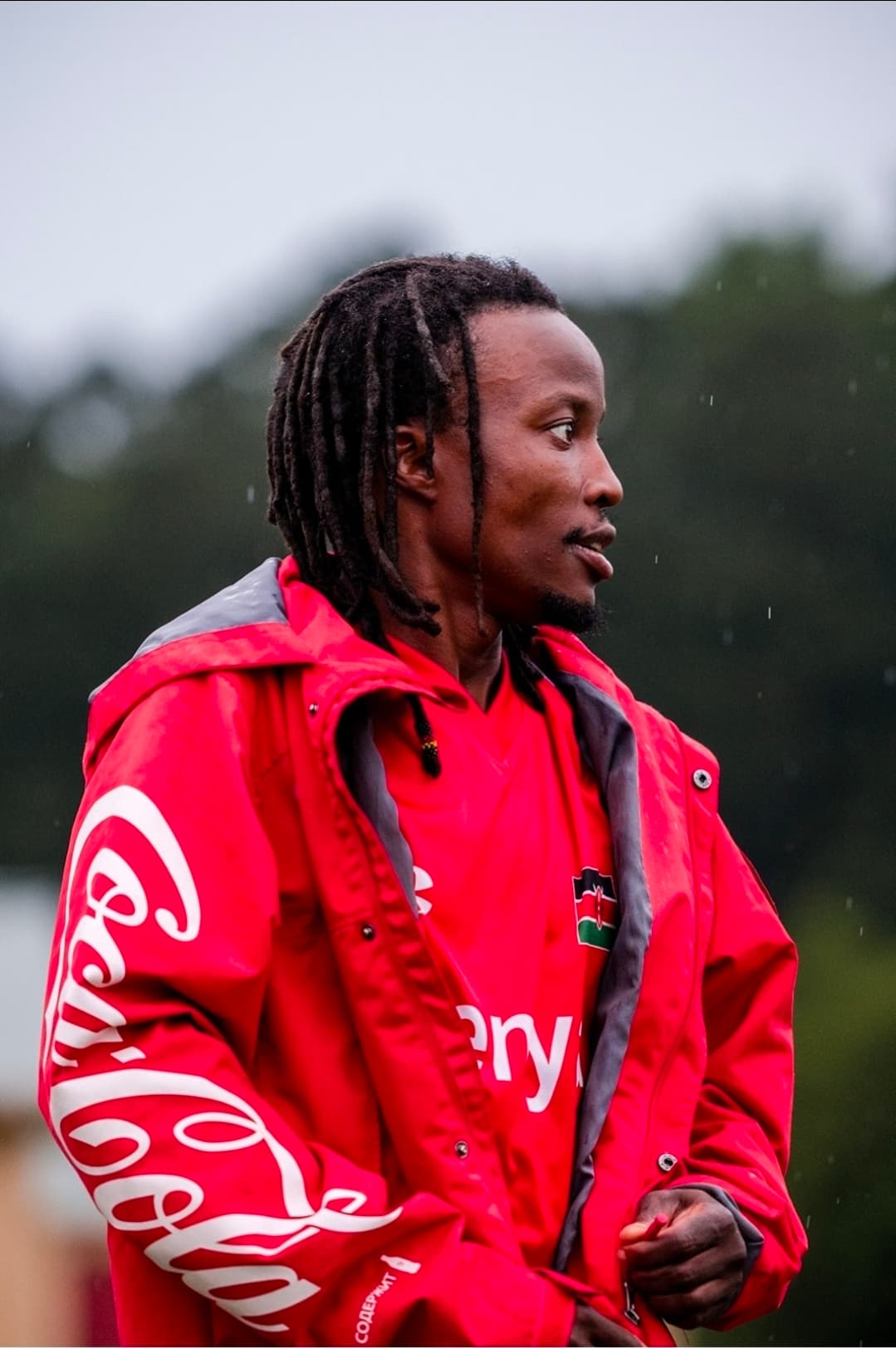 A JFC staff member in a red jacket and Kenya shirt looking out across the pitch in the rain