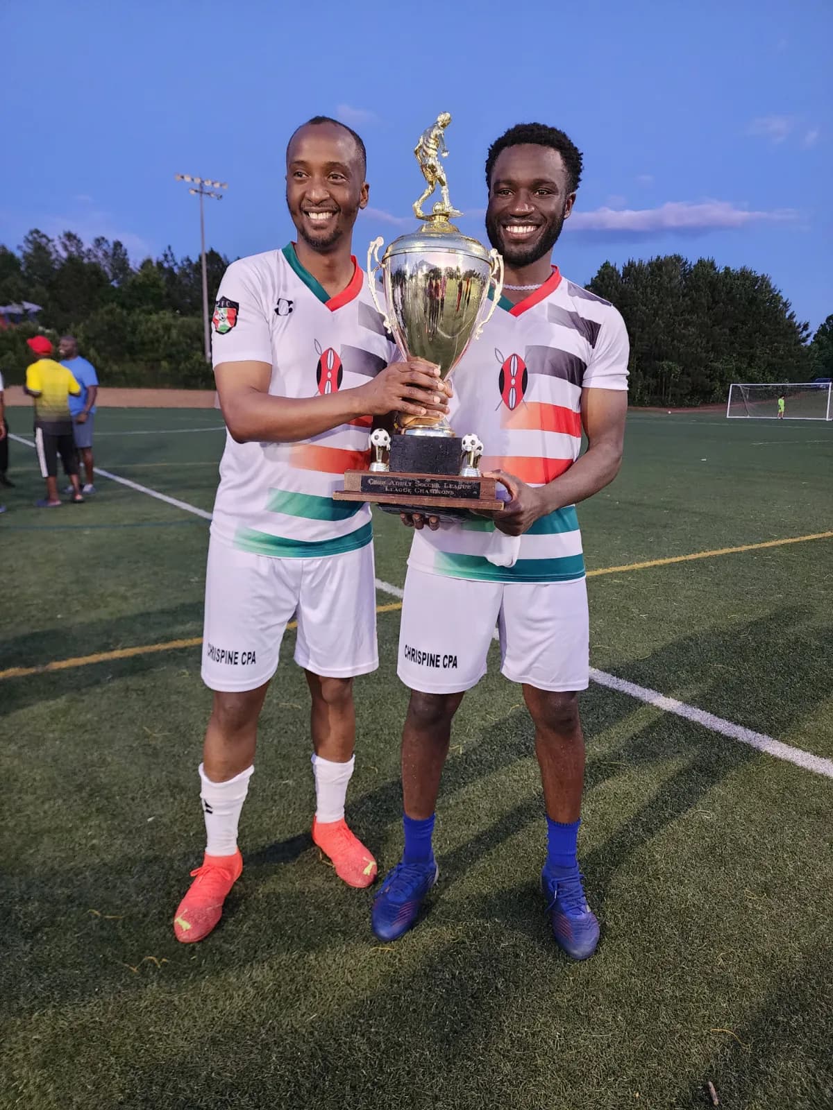 Two players in white striped kit holding a trophy together at dusk