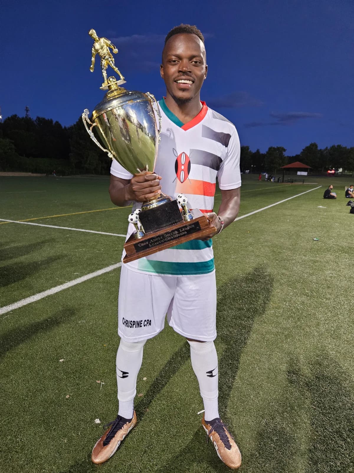 A player in white striped kit lifting a trophy on the pitch at dusk