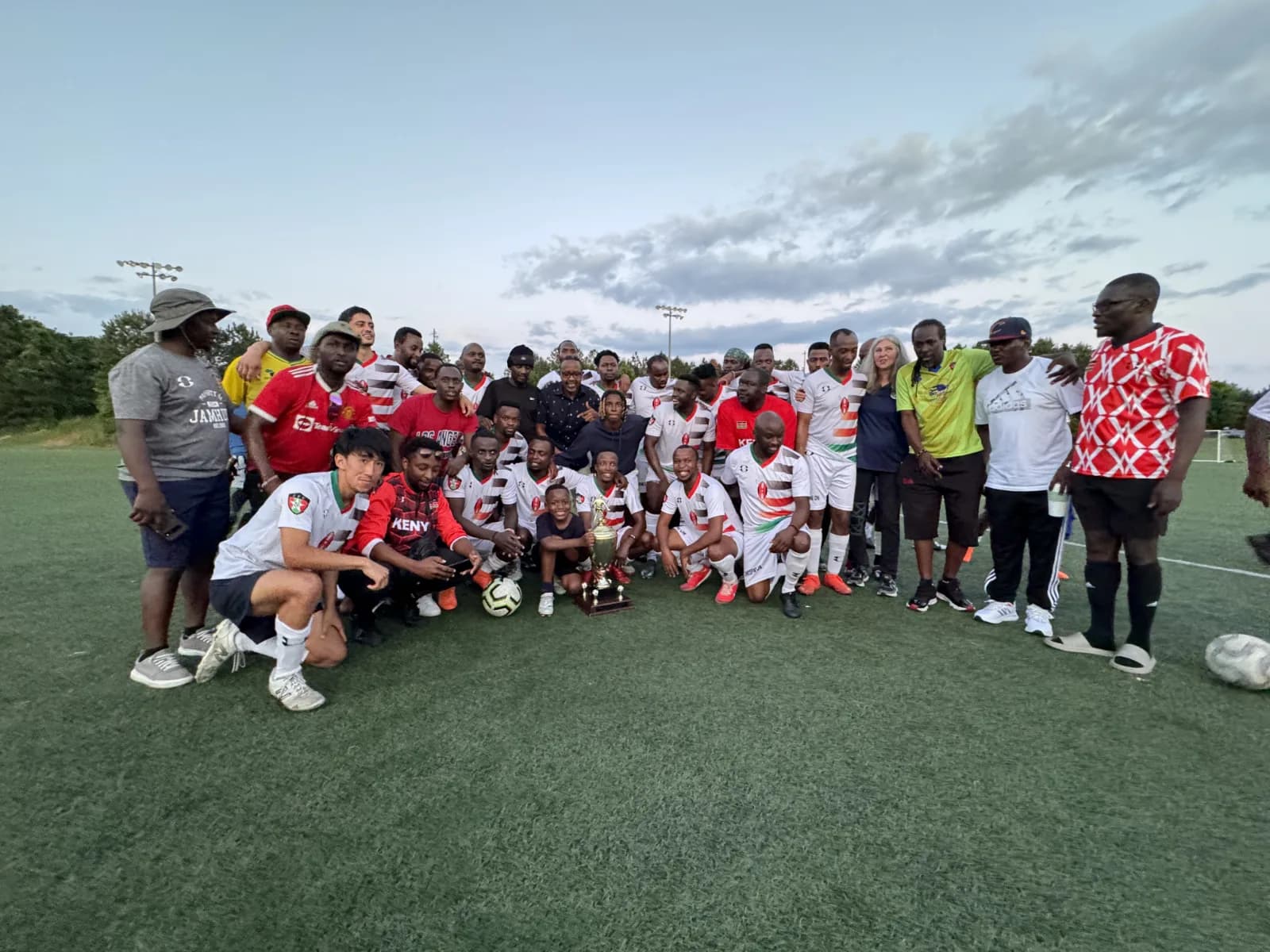 A wide group photo of players and supporters on the pitch at dusk