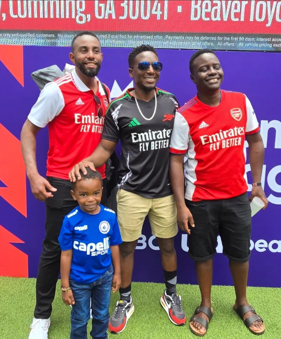 Three men in Arsenal shirts with a young boy in blue in front of a Premier League backdrop