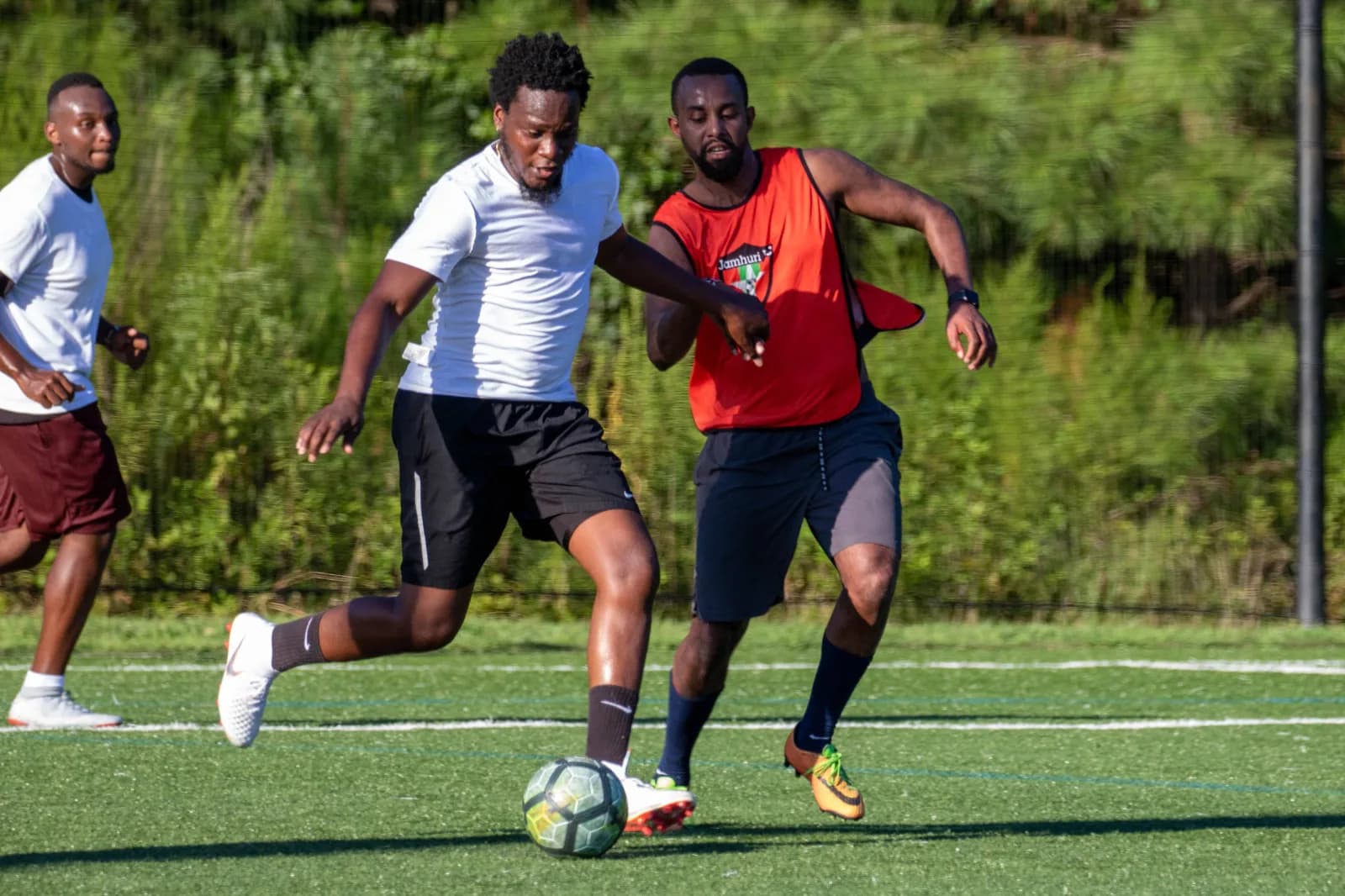 A player in a white shirt and a player in an orange bib chase the ball