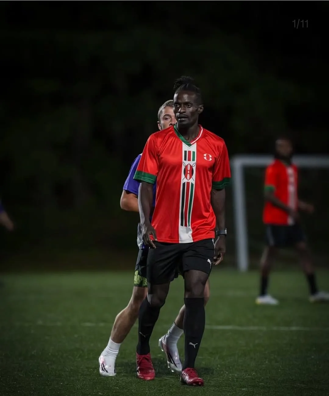 A JFC player in red standing on the floodlit pitch, eyes on the play